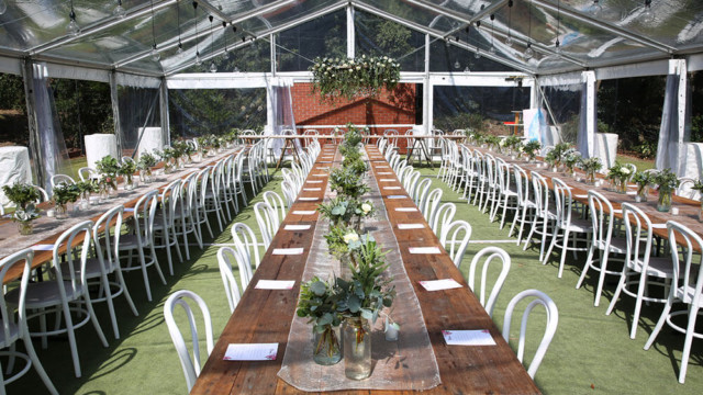 A long table surrounded by white chairs, adorned with green plants, set under garden marquees.
