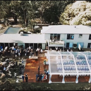 Aerial view of a wedding party gathered in front of a house, with white wedding marquees set up on the lawn.