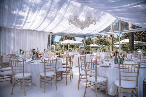 Elegant marquee wedding setup with white drapery, chandeliers, and beautifully arranged dining tables surrounded by lush greenery in an outdoor setting which will influence the average costs of a marquee wedding.