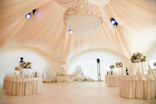 Luxurious marquee wedding interior featuring peach drapes, a grand floral chandelier, and elegant round tables with floral centrepieces under soft lighting which can influence the marquee hire costs.