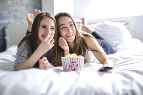 Two teenage girls laughing and eating popcorn during a movie night.